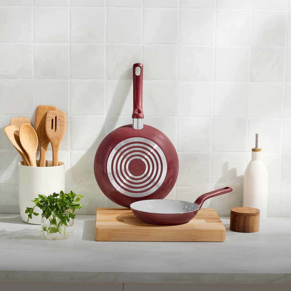 Large red frypan standing upright behind smaller red frypan on wooden board