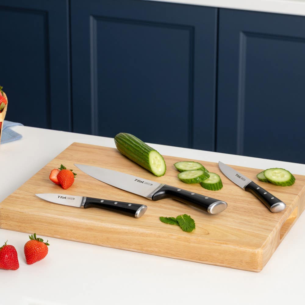Three T-fal knives on a cutting board with sliced cucumber and strawberries.