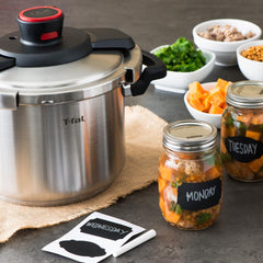 A stainless steel T-fal pressure cooker with black handles on a kitchen counter near labeled jars labeled, and food ingredients.