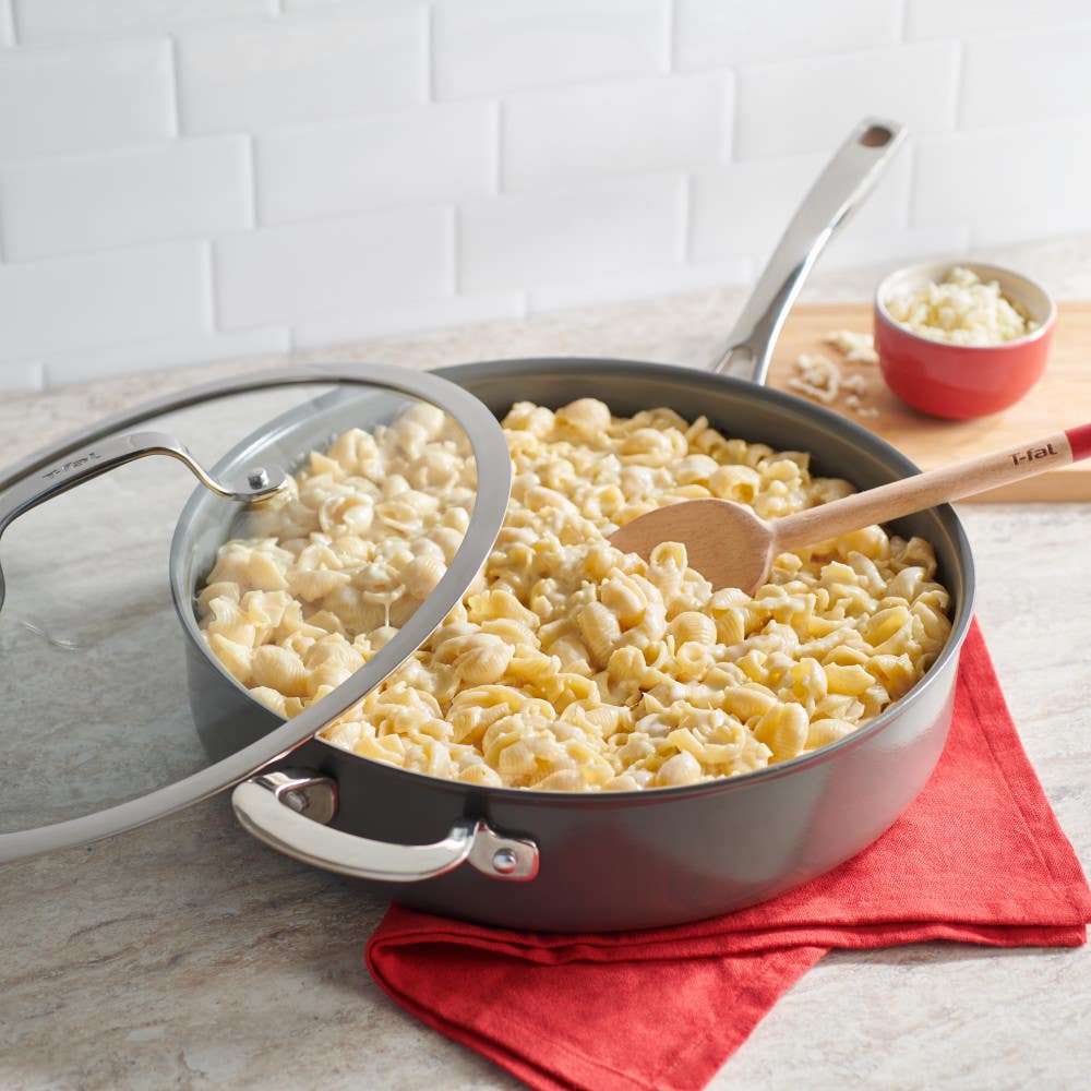 A pan of pasta with a wooden spoon, placed on a red cloth with a small bowl of cheese nearby.