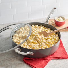 A pan of pasta with a wooden spoon, placed on a red cloth with a small bowl of cheese nearby.