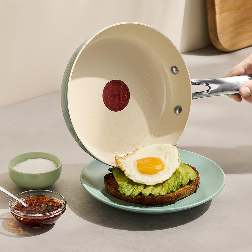 A fried egg slides from a ceramic skillet onto avocado toast on a plate.