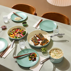 Set table with pastel plates, glasses, chopsticks, and pans of rice, salmon, and vegetables.