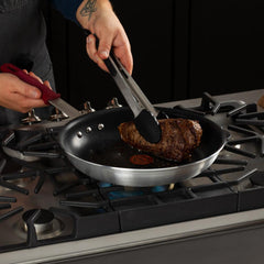 Hand turning browned steak with tongs in t-fal nonstick frypan over a gass stovetop with blue gas flame