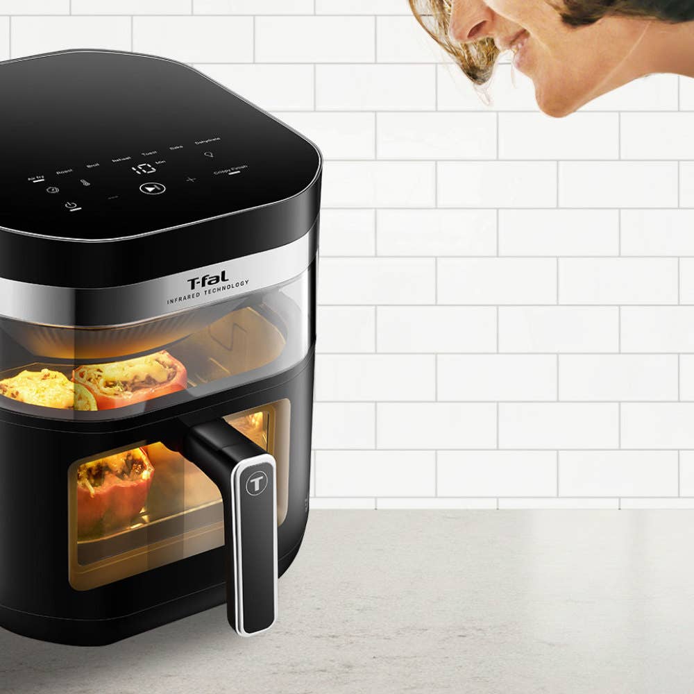 A woman looking at an air fryer through two viewing windows, with colorful vegetables cooking inside.