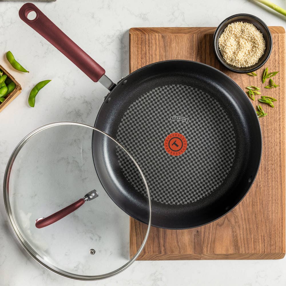 A frying pan with red handle and T-fal thermospot heat indicator on a wooden board beside ingredients and a glass lid.