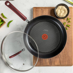 A frying pan with red handle and T-fal thermospot heat indicator on a wooden board beside ingredients and a glass lid.