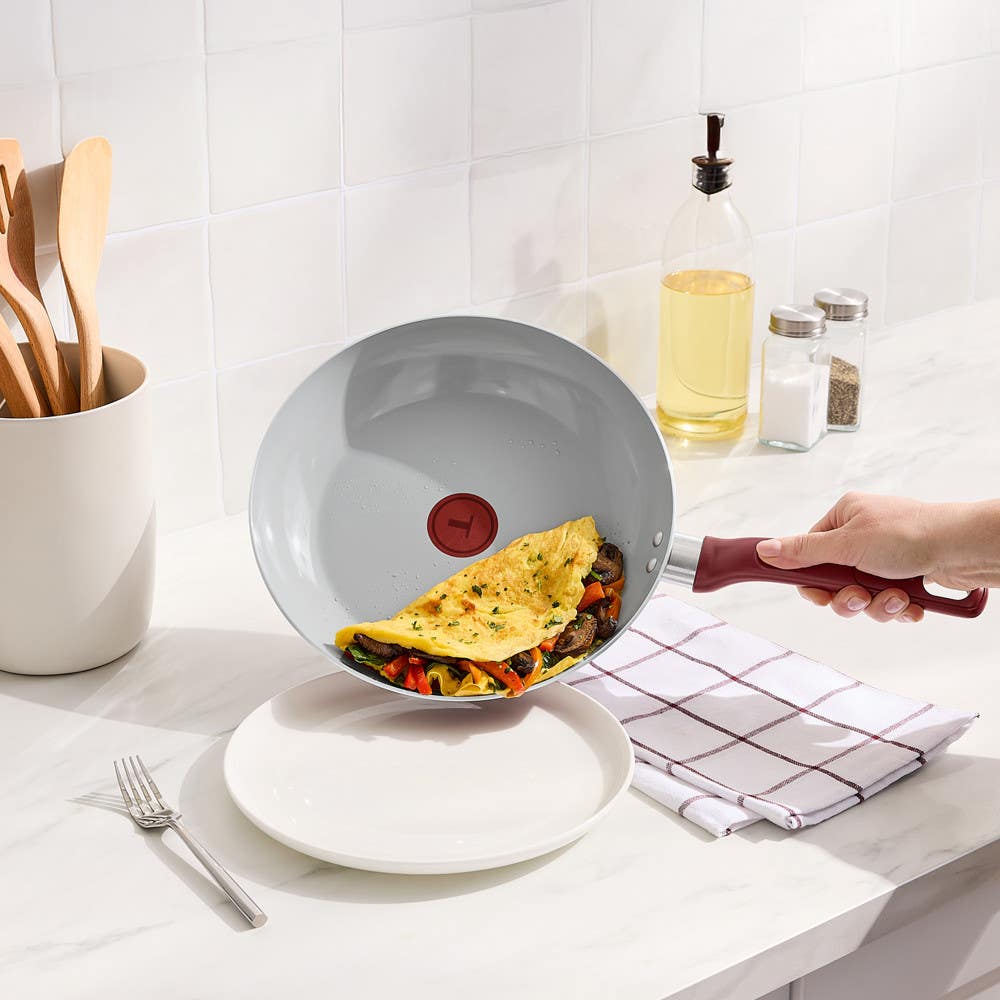 Hand tilting red frypan to serve vegetable omelet onto white plate on countertop