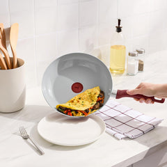 Hand tilting red frypan to serve vegetable omelet onto white plate on countertop