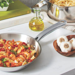 Stainless steel frypan with pasta, tomatoes, and parsley displayed next to garlic and olive oil.