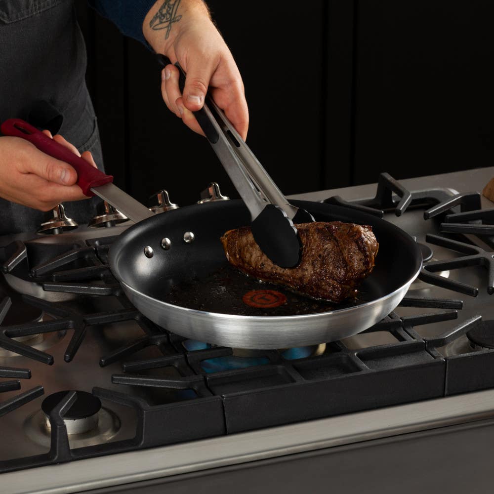 A person is searing a piece of steak in a nonstick frying pan over a gas stove using tongs