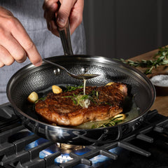 A person cooking a steak and basting it with herbs and garlic in a frypan on a stovetop
