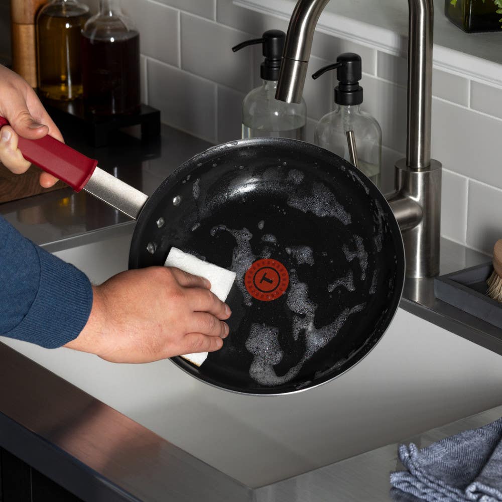 A person washes a black interior nonstick frying pan with soap and water in a kitchen sink