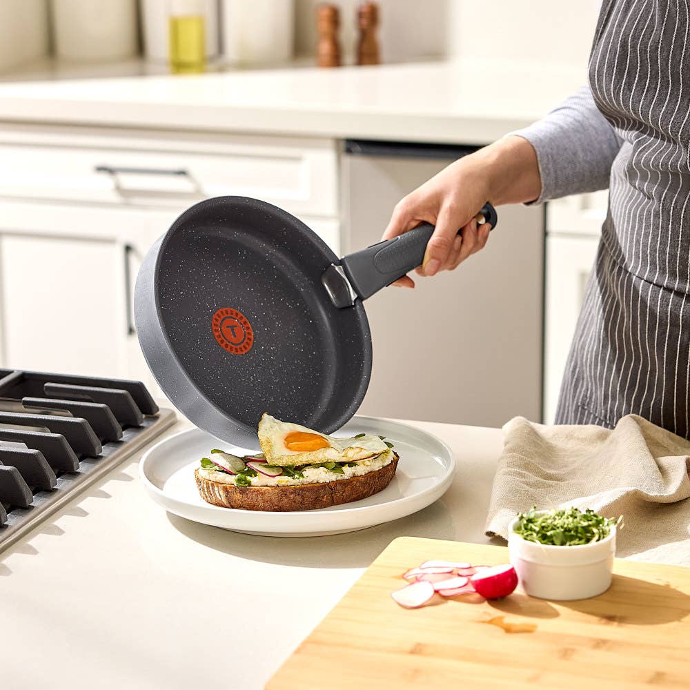 Hand sliding a fried egg from a gray pan with detachable handle onto toast with avocado