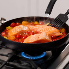 A person uses a black spatula to cook three salmon fillets and cherry tomatoes in a black nonstick frying pan on a stovetop.